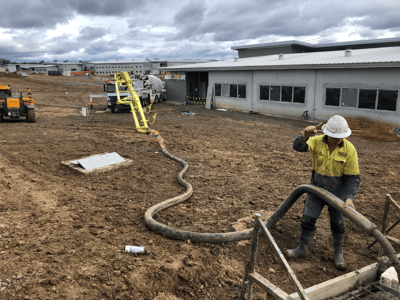 A construction worker in a high-vis shirt and hard hat guiding a flexible concrete hose on a muddy construction site, connected to a distant yellow pump truck.
