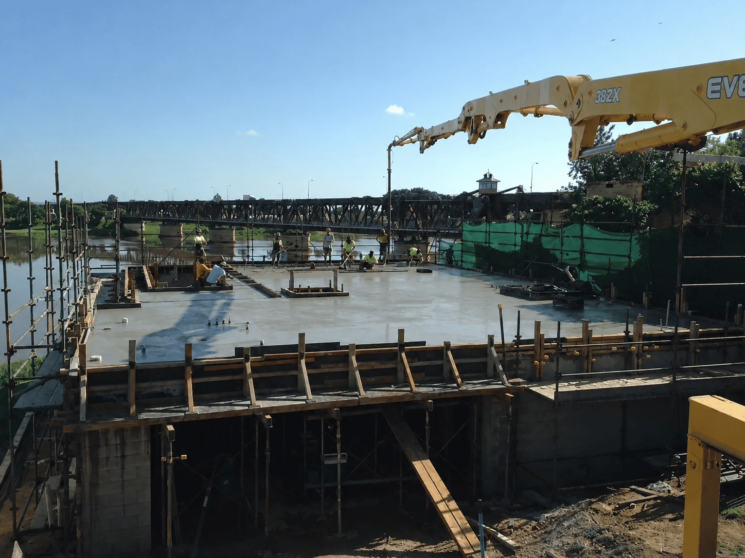 A yellow concrete pump arm extending over a elevated construction site near a river and bridge as workers smooth fresh concrete.