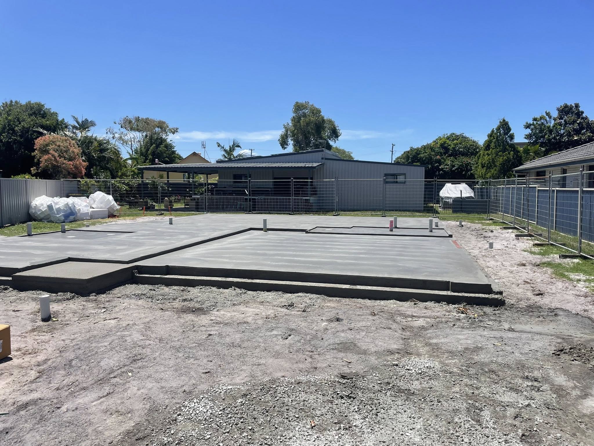 A smooth, freshly poured concrete foundation slab surrounded by temporary construction fencing on a sunny day.