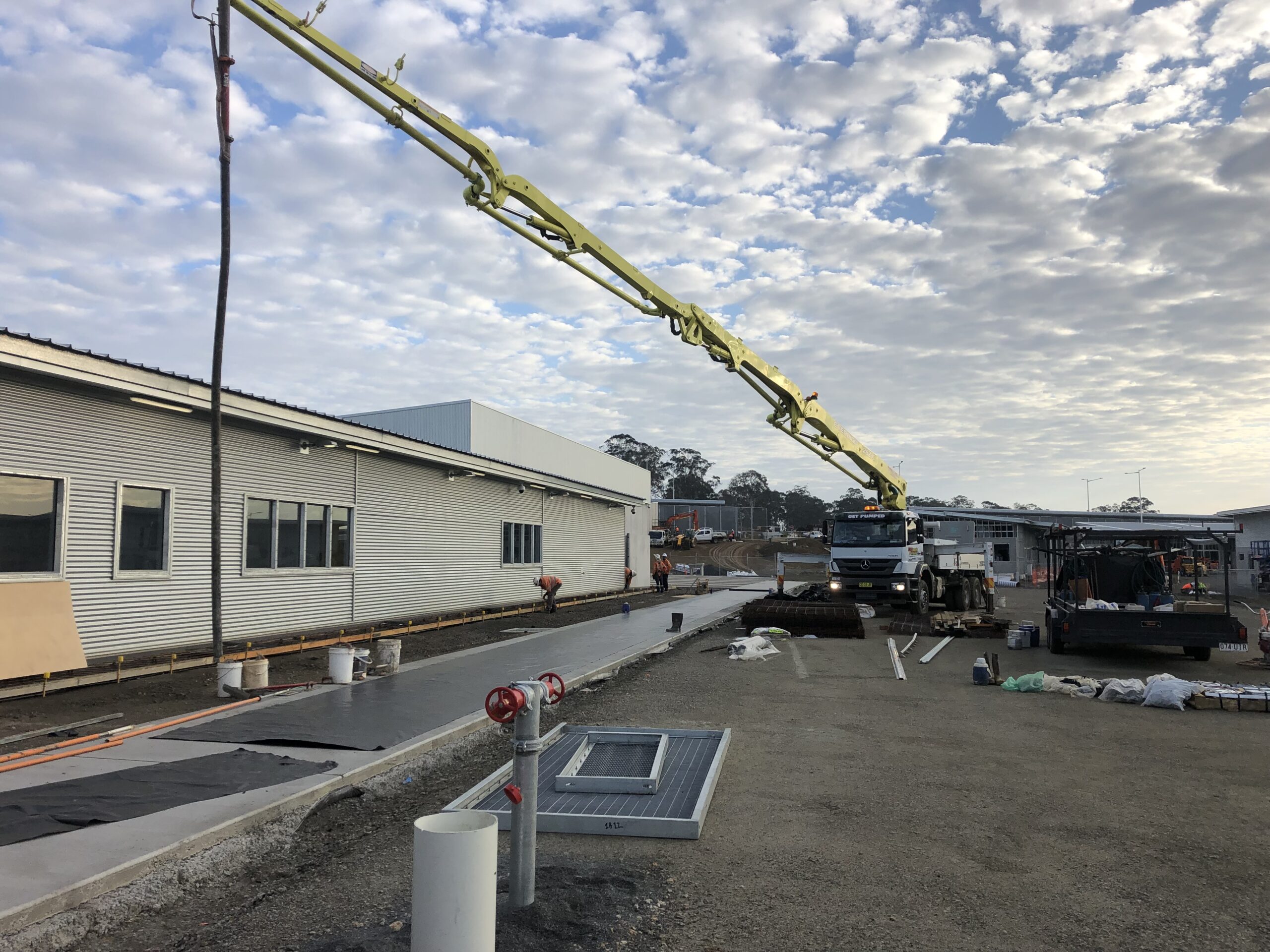 A yellow concrete pump truck extending its long boom to pour concrete for a walkway beside a building.