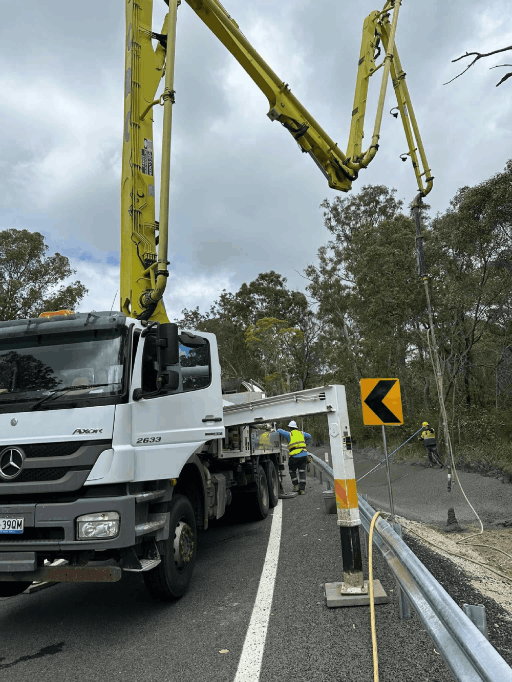 A white concrete pump truck parked on a curved road, extending its yellow boom over a guardrail to pour concrete along the roadside.