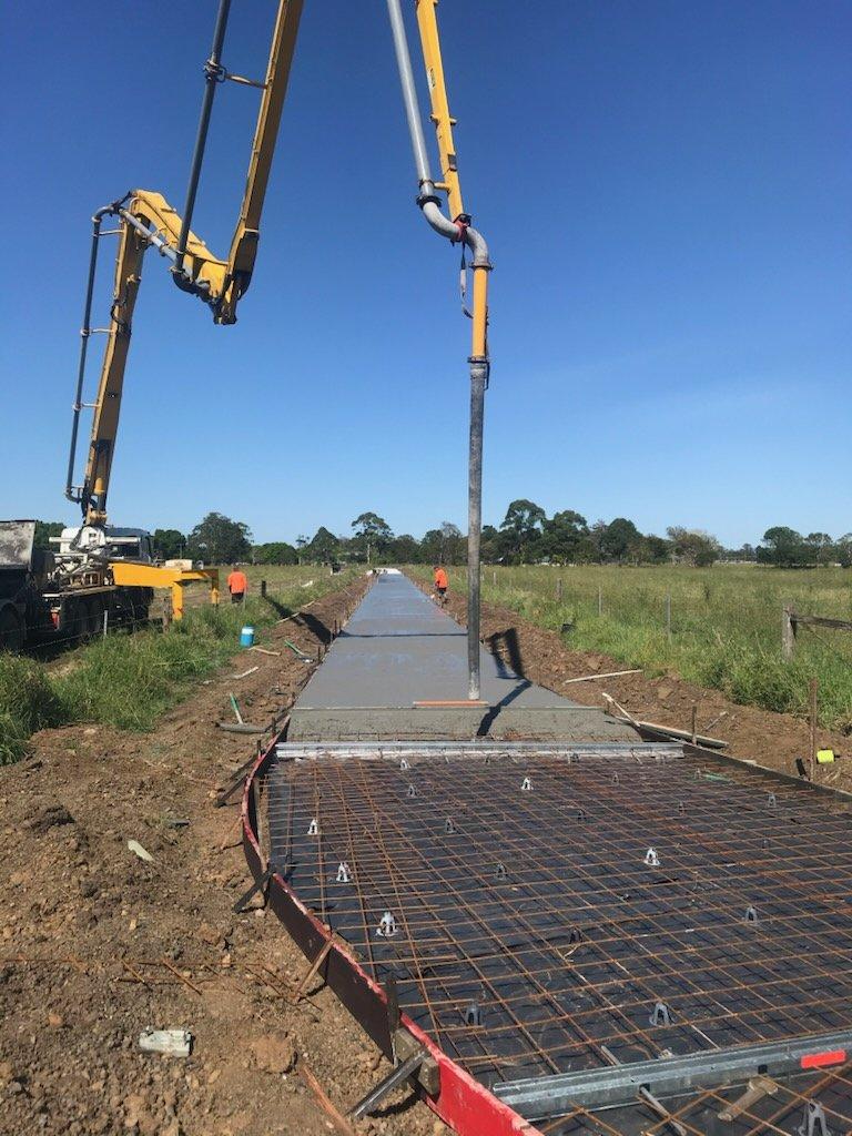 A yellow concrete pump truck extending its long boom to pour a straight concrete pathway through a grassy rural field.