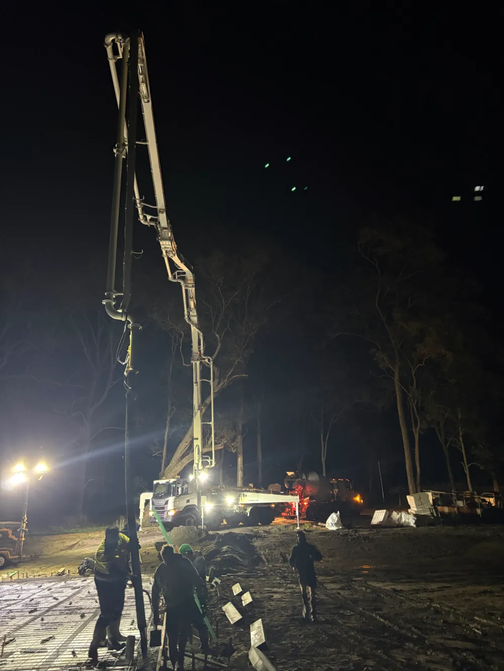 A white concrete pump truck with an illuminated, fully extended boom operating on a construction site at night.
