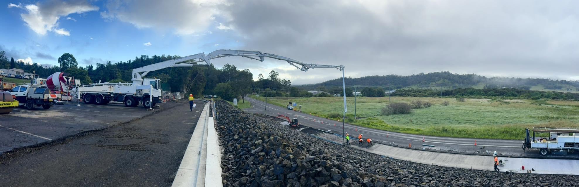 Panoramic view of a white concrete pump truck with an extended boom operating along a rock embankment on a rural road project.
