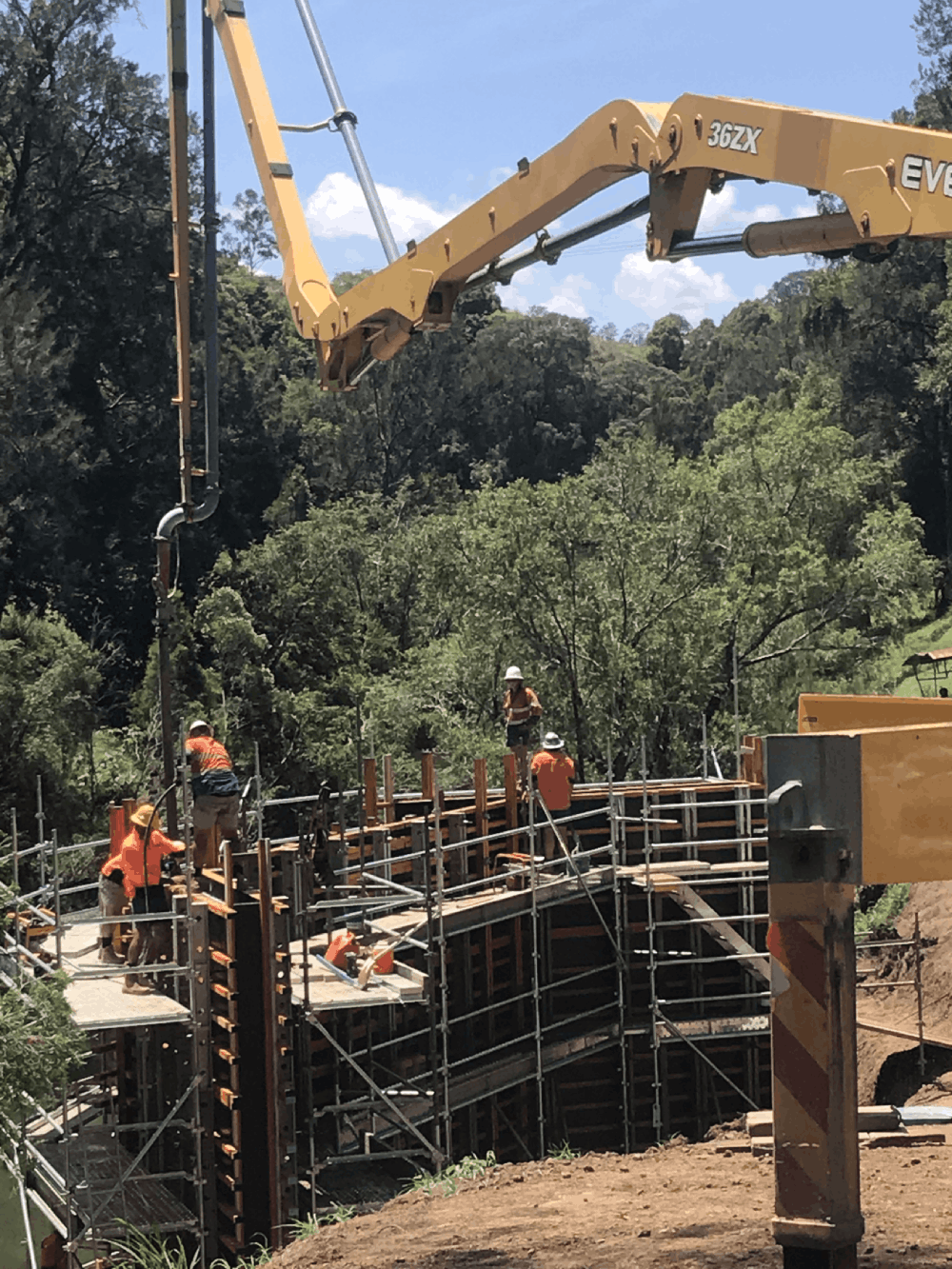 A yellow concrete pump boom extending over construction scaffolding where workers in high-vis shirts manage a pour in a wooded area.