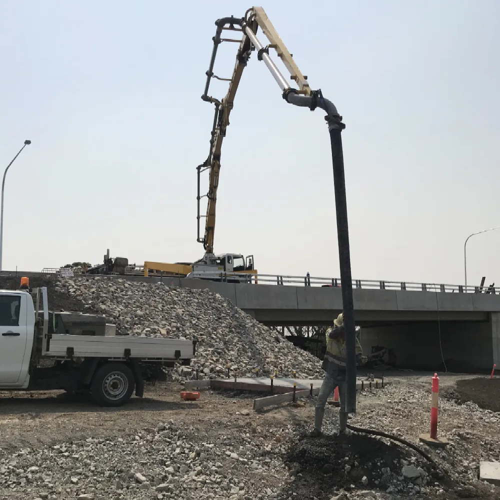 A construction worker guiding a concrete pump hose near a bridge overpass with a pump truck parked on the upper road level.